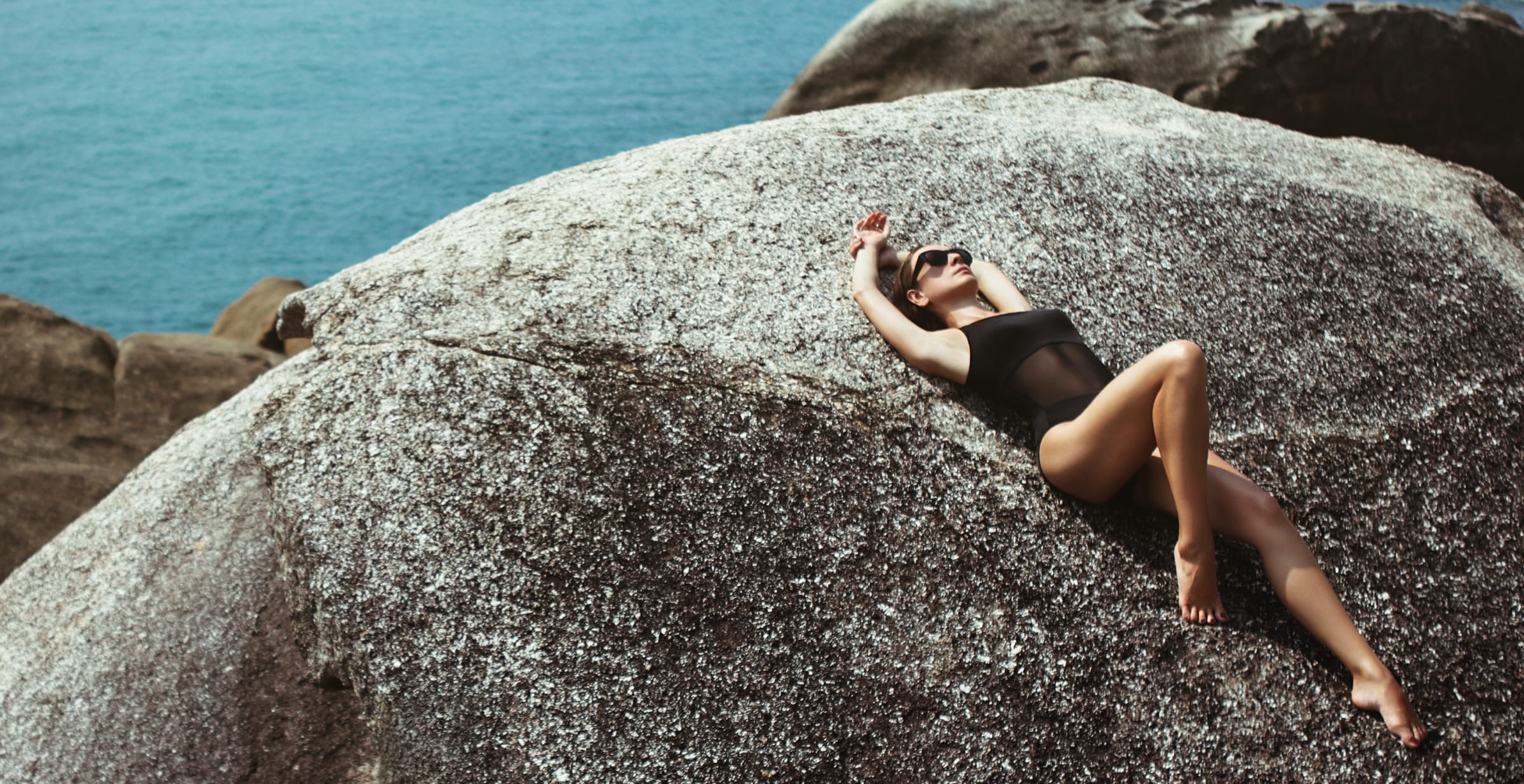A woman in a black swimsuit resting on a sunlit rock above the sea